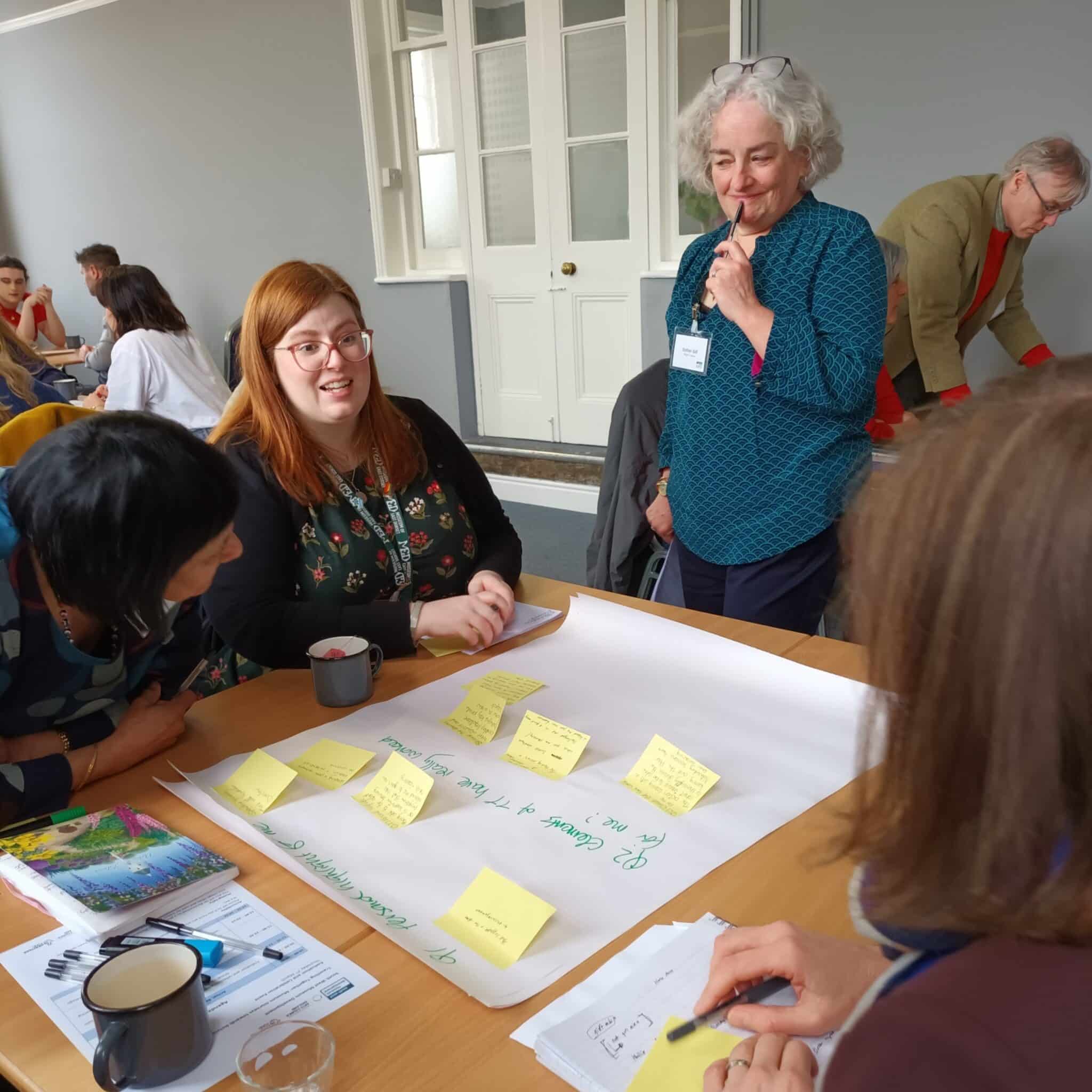 A woman stands looking at a table. On the table there is a large piece of paper with post-it notes on it. Around the table three people are animatedly discussing something. 