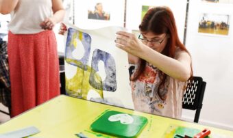 A woman sits at a yellow table; she holds up a print that she has made.